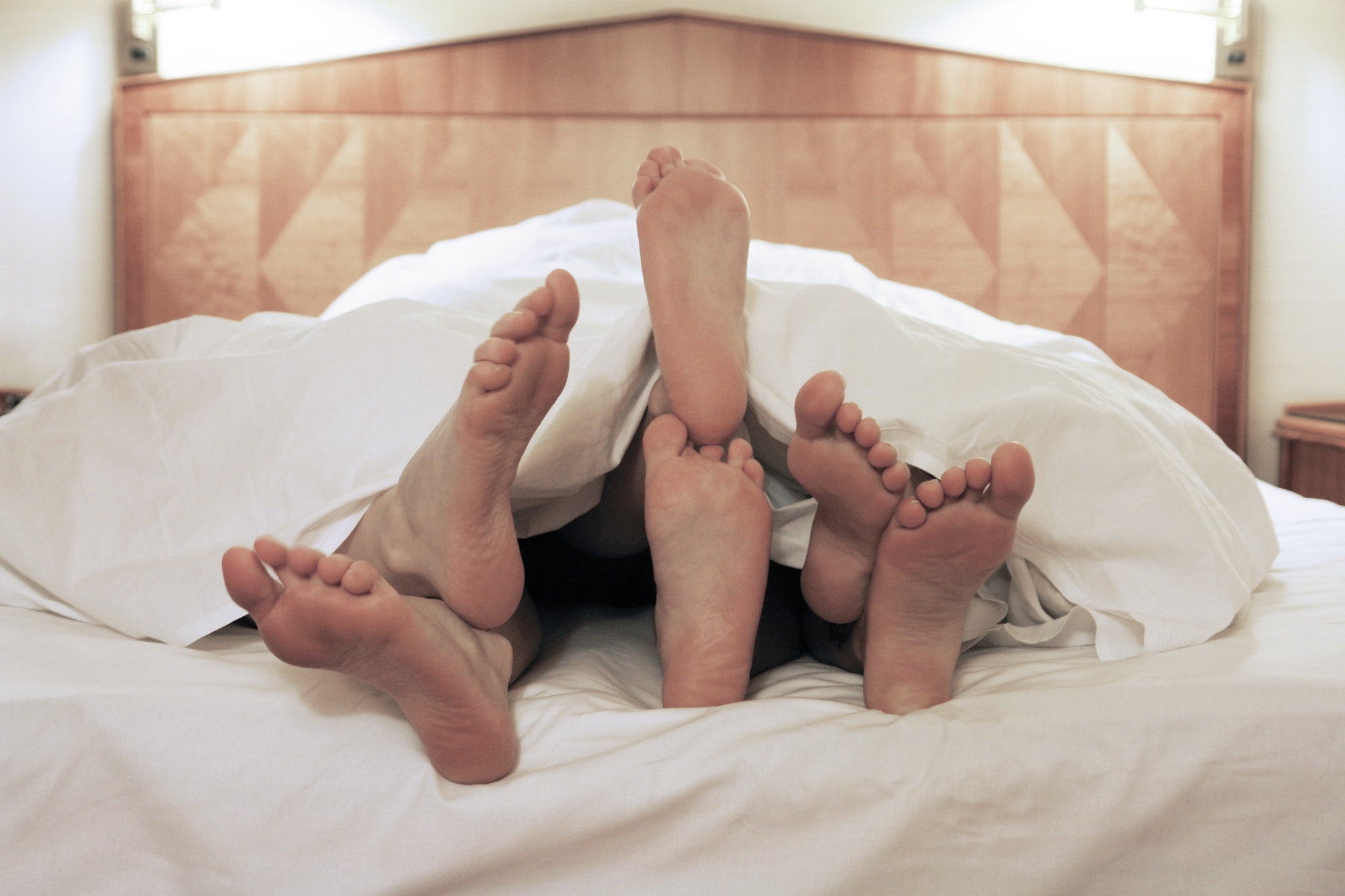 three unrecognizable people lying in one bed together. feet showing under duvet. lesbian threesome